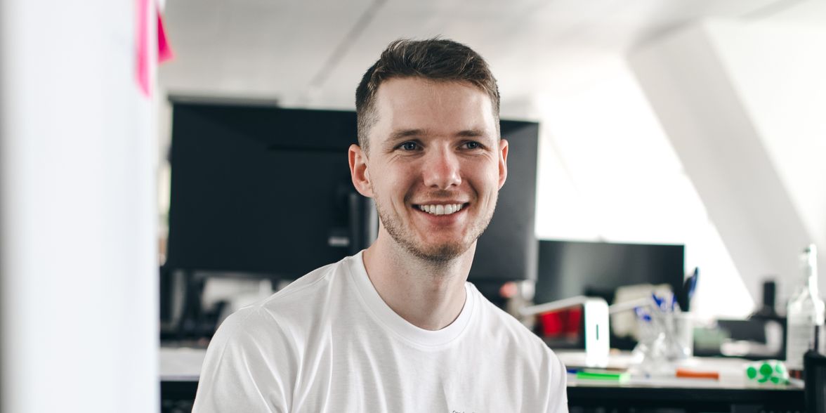 Smiling man in a modern office, wearing a white shirt with English text, testimonial setting.