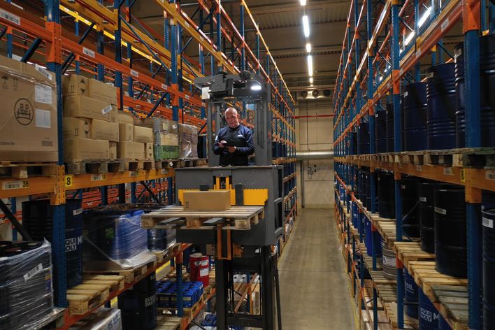 Warehouse worker operates lift among shelves with boxes and barrels; no software UI or keywords visible.