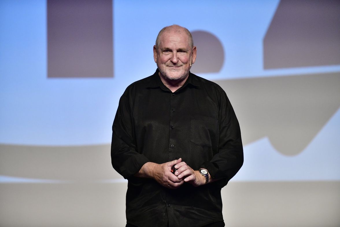 Man in black shirt standing on stage with abstract background, no visible text or software UI.
