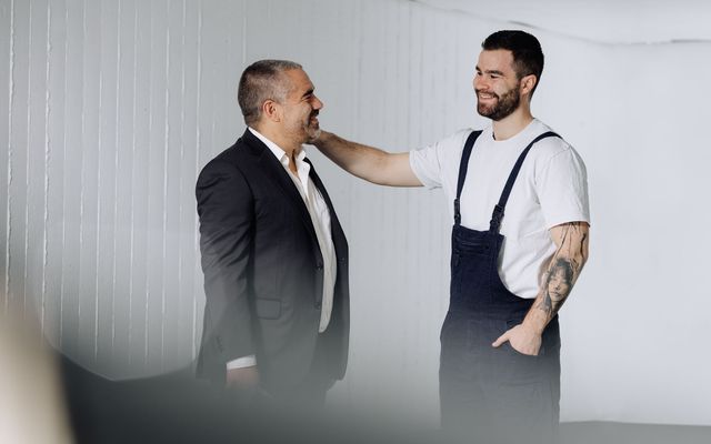 Two men, one in a suit and one in work overalls, smiling and interacting in a bright room.