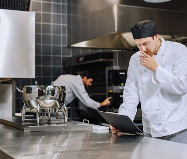 Two chefs working in a professional kitchen, one reading a clipboard, stainless steel equipment visible.