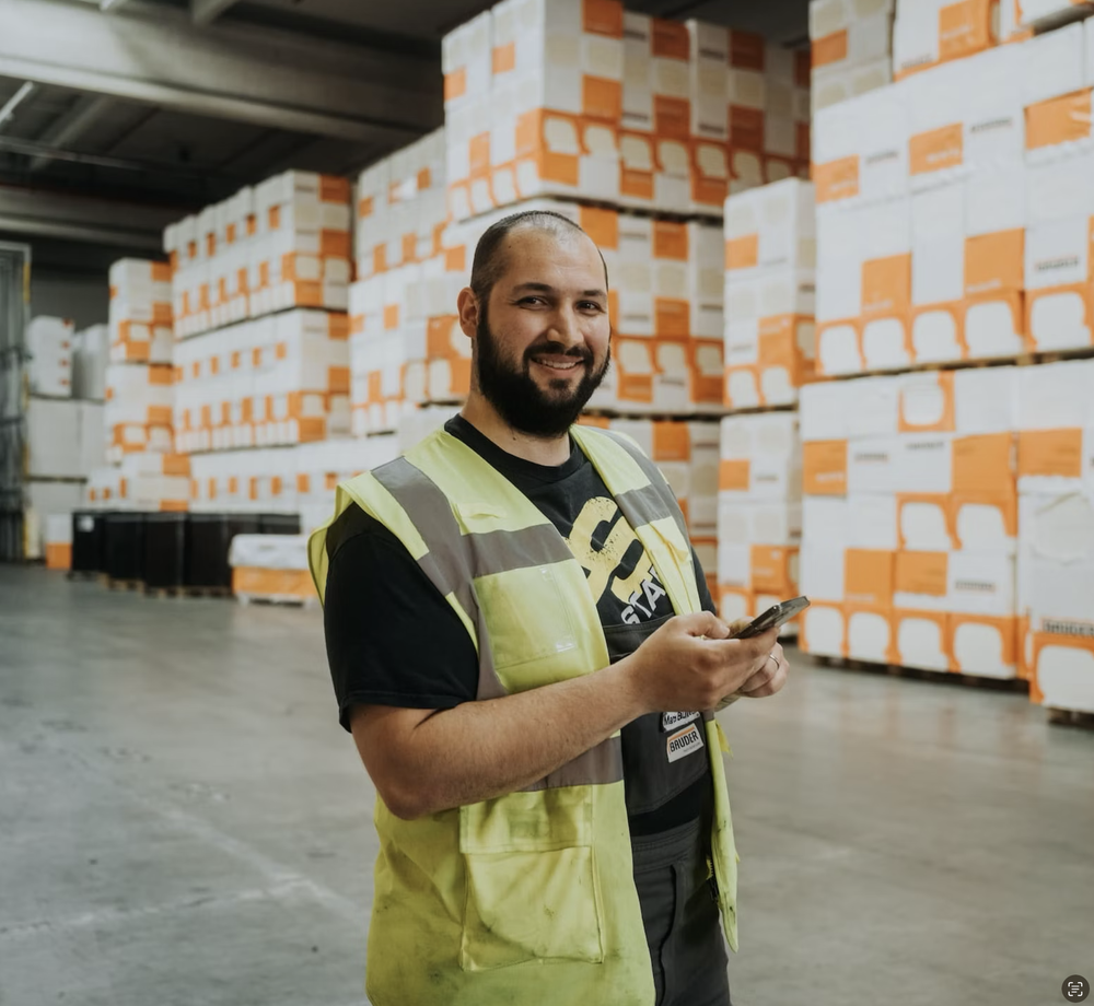 Warehouse worker in a safety vest using a smartphone among Bauder boxes.