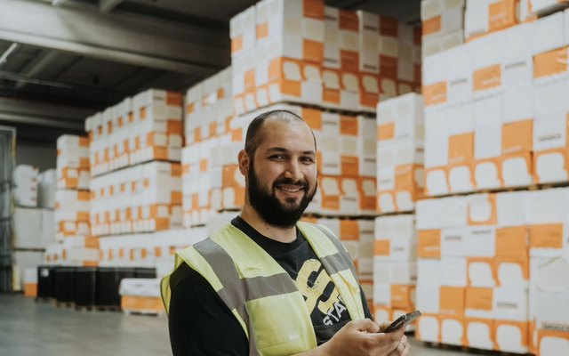 Warehouse worker in safety vest using smartphone, surrounded by stacked Bauder packaging boxes.