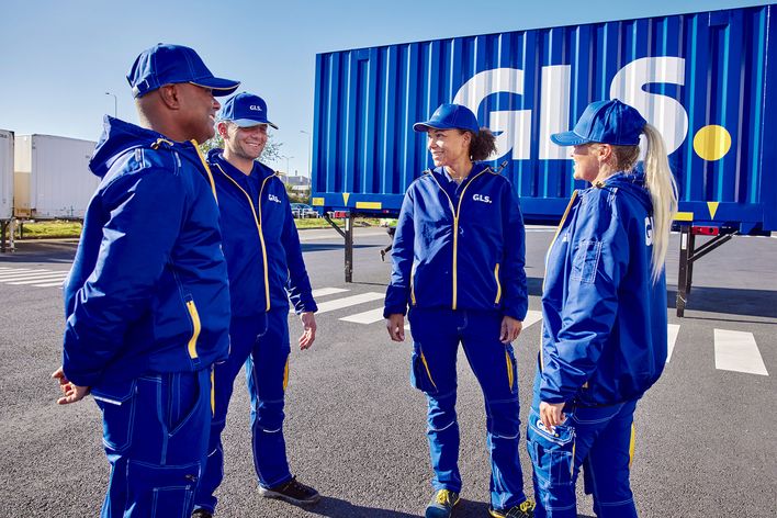 Four GLS employees in blue uniforms stand and talk outside near a GLS truck container.