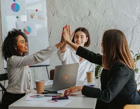 Three businesswomen high-five at a desk with charts, symbolizing teamwork and employee engagement.