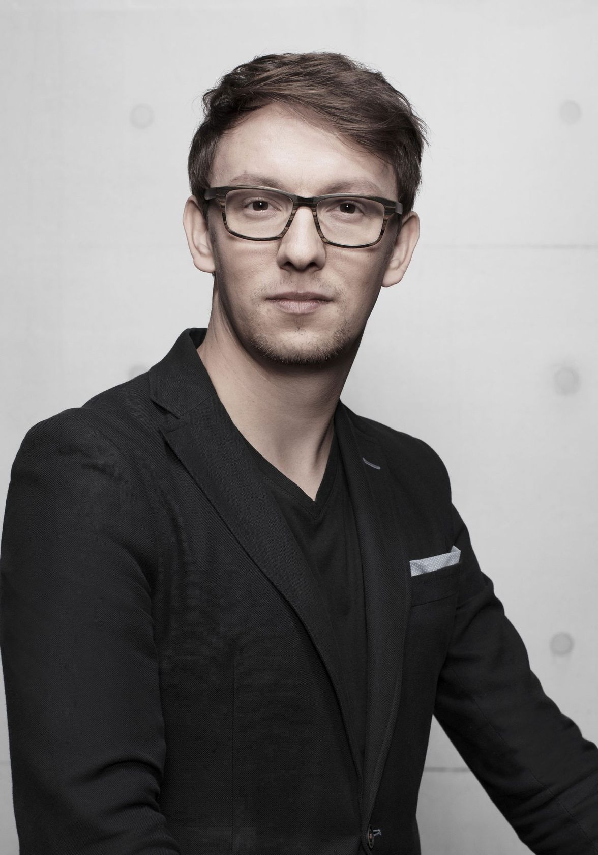 A person in glasses and a black blazer against a plain gray background.