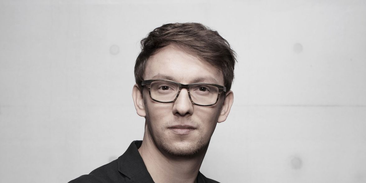 A person in glasses and a black blazer against a plain gray background.