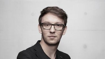 Portrait of a man in a black suit and glasses against a plain, light background.