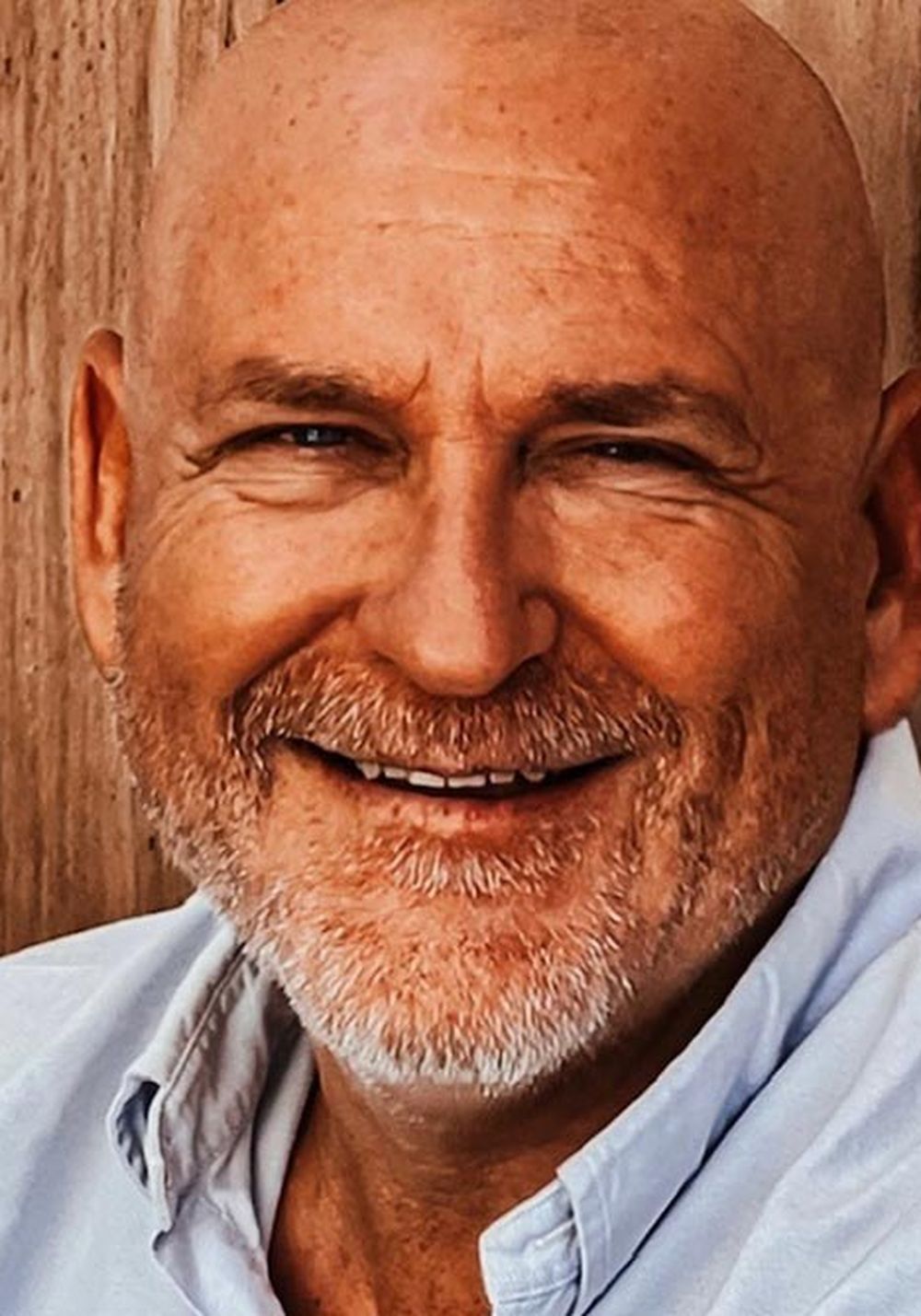 Portrait of a smiling bald man with a beard in a light shirt, sitting in front of wooden panels.