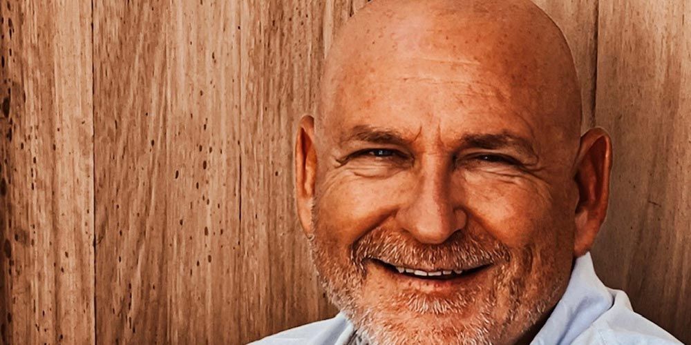 Portrait of a smiling bald man with a beard in a light shirt, sitting in front of wooden panels.