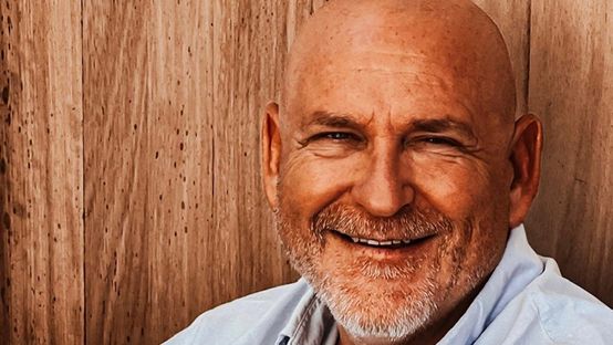Portrait of a smiling bald man with a beard in a light shirt, sitting in front of wooden panels.