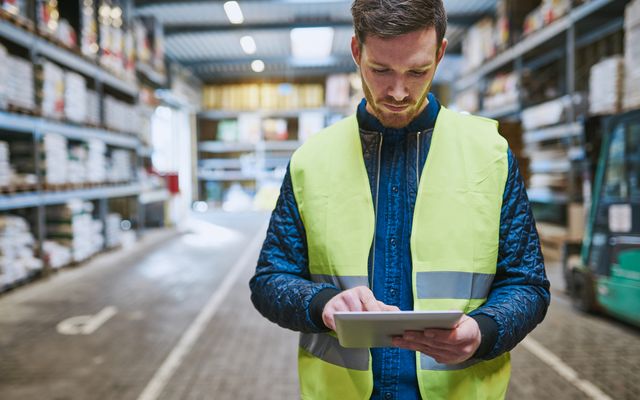 Warehouse worker in safety vest uses tablet, representing new work for blue-collar frontline staff.
