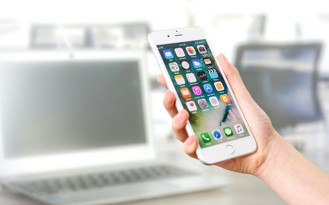Hand hält Smartphone vor Laptop, Fokus auf Apps, modernes Büro, digitale Mitarbeiterplattform.