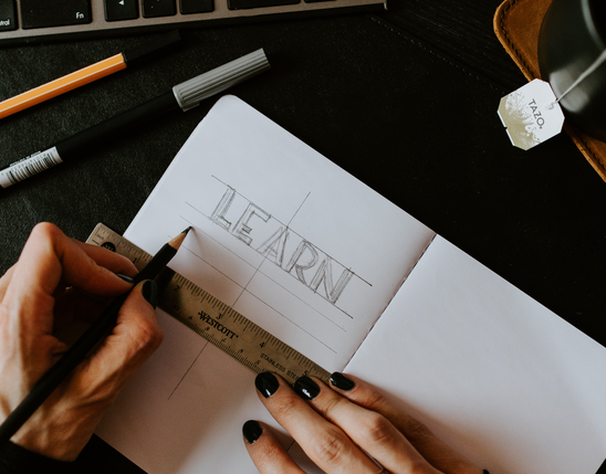 A person sketches LEARN in a notebook with a ruler, near a keyboard, pens, and tea.