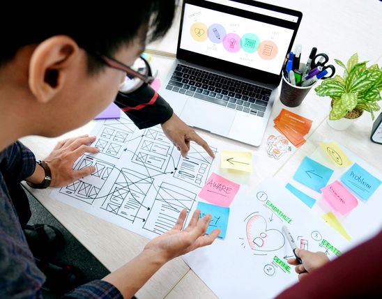 People collaborating on UX design with wireframes, sticky notes, and a laptop showing a workflow diagram.