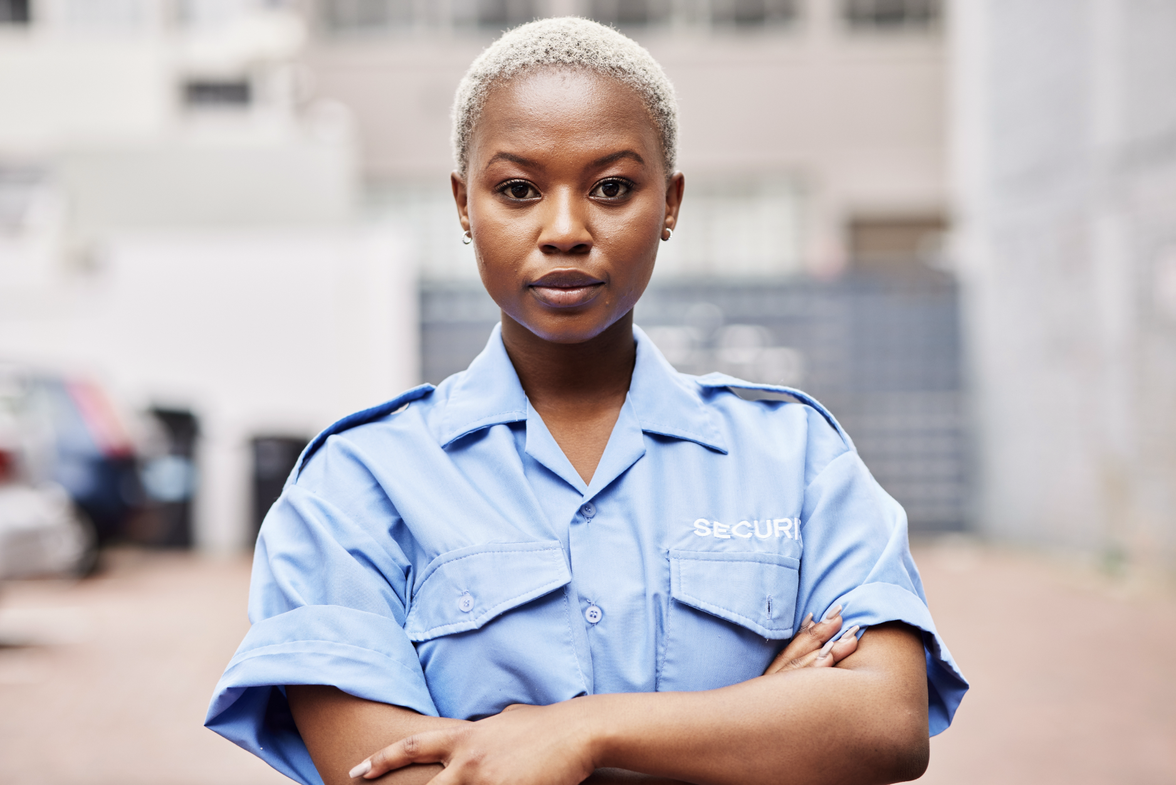 Security guard in blue uniform with SECURI text, standing outdoors with arms crossed.