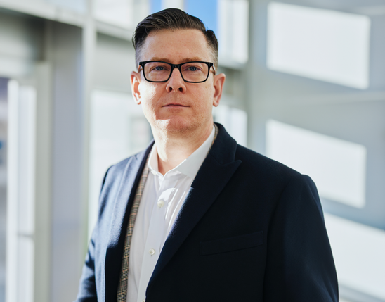 Man standing beside a wall in a sunlit modern office environment, wearing glasses and a suit.