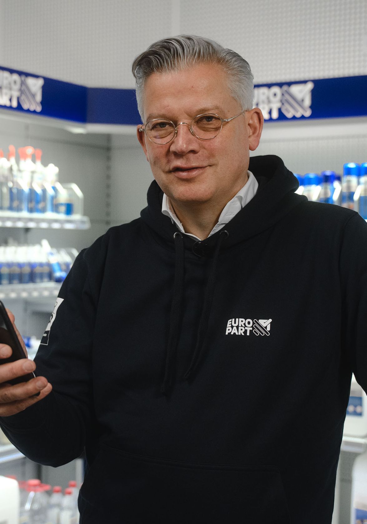 Man in EURO PART hoodie holding smartphone in store aisle with shelves of automotive products.