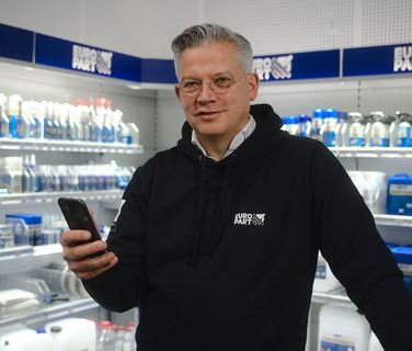 Man in EURO PART hoodie holding smartphone in store aisle with shelves of automotive products.