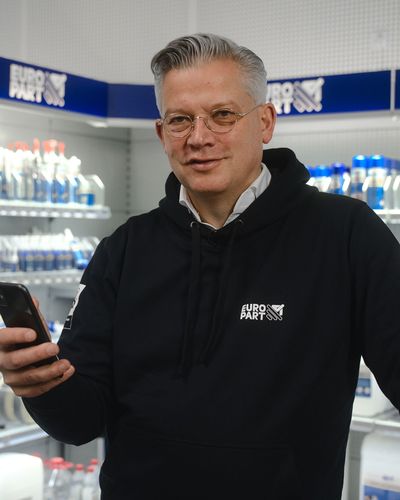 Man in EURO PART hoodie holding smartphone in store aisle with shelves of automotive products.