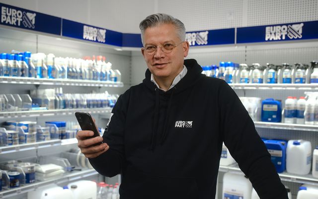 Man in EURO PART hoodie holding smartphone in store aisle with shelves of automotive products.