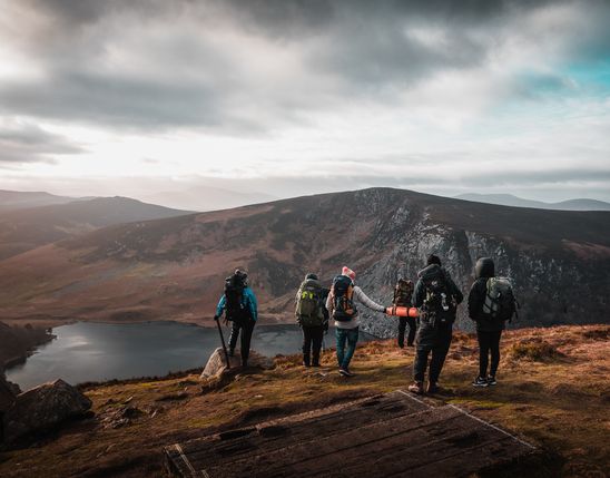A group of hikers with backpacks stands on a mountain summit overlooking a lake and valleys in Ireland.