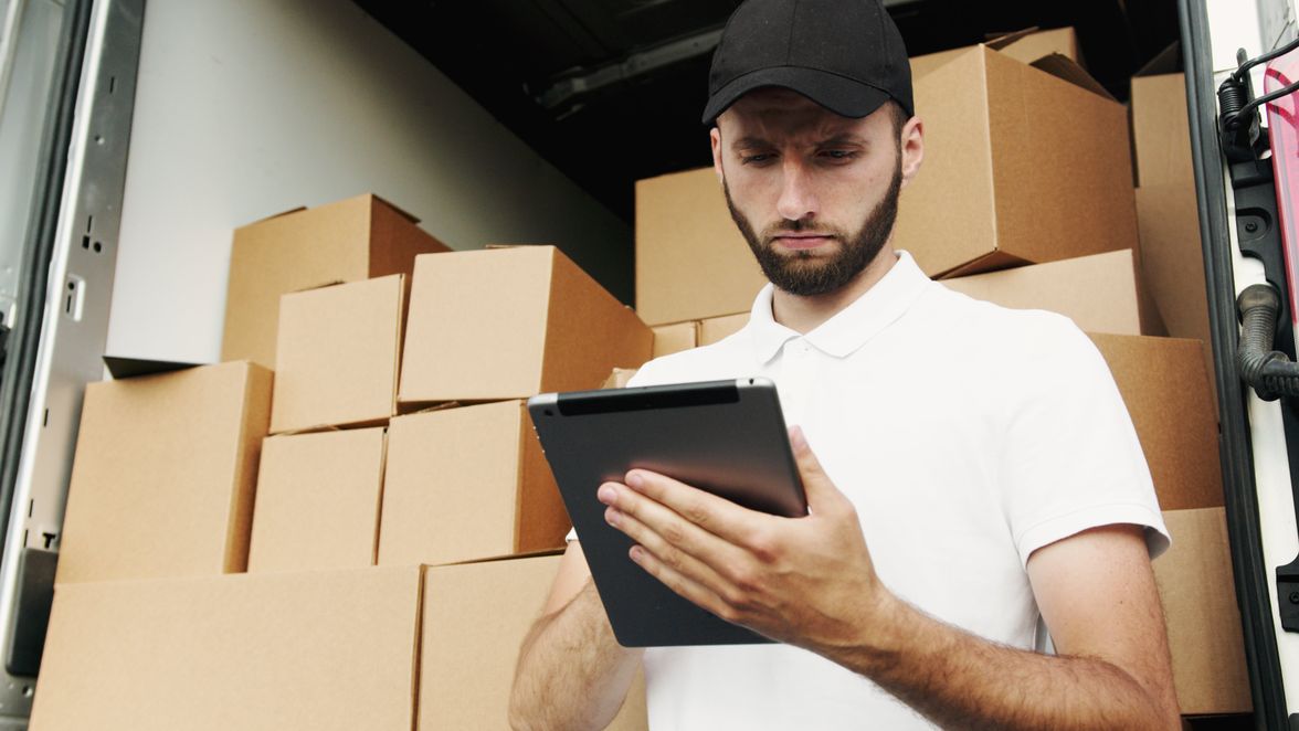 Man in a cap using a tablet in front of stacked cardboard boxes.