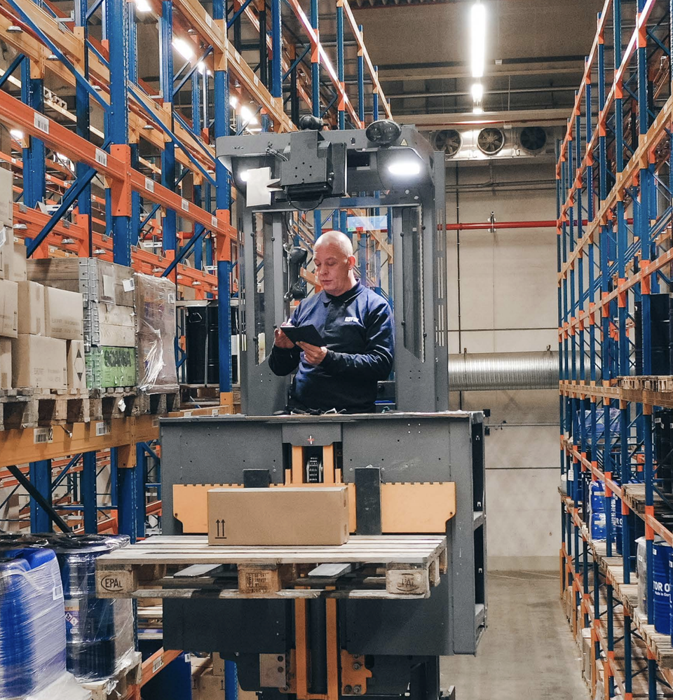 Warehouse worker operating a lift, handling packages among shelves, no visible software interface.