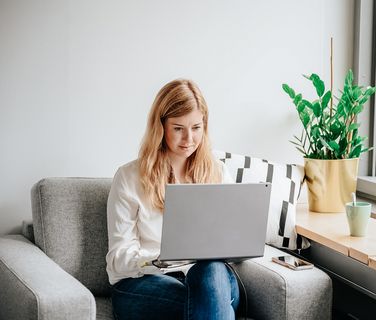 Woman working remotely on a laptop in a home office with a plant and coffee mug nearby.