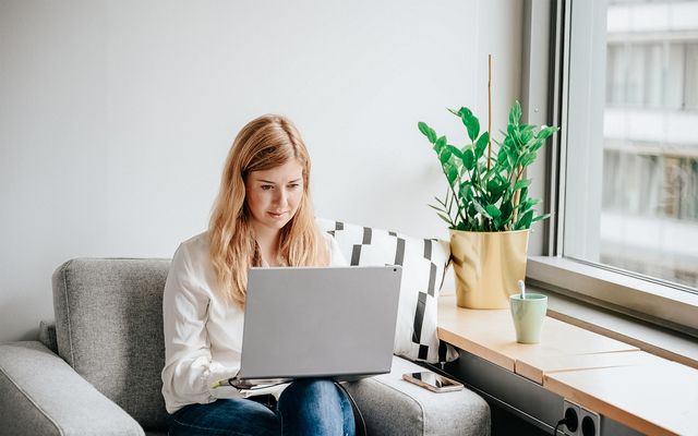 Woman working remotely on a laptop in a home office with a plant and coffee mug nearby.