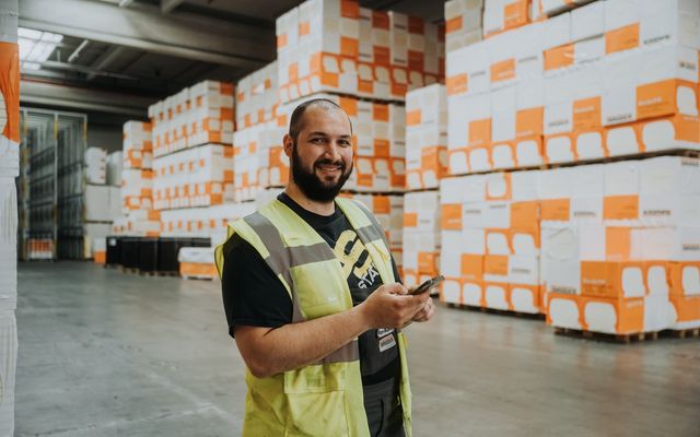 Warehouse worker in safety vest using smartphone, surrounded by Bauder-labeled pallets.