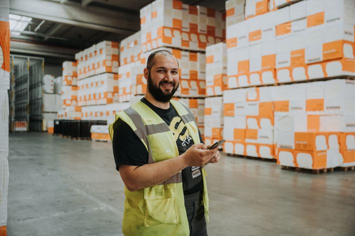 Warehouse worker in safety vest using smartphone, surrounded by Bauder-labeled pallets.