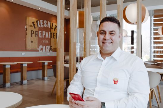 McDonalds employee smiling in restaurant, holding phone, FRESHLY PREPARED WITH CARE on wall.