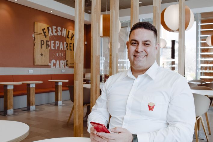 McDonalds employee smiling in restaurant, holding phone, FRESHLY PREPARED WITH CARE on wall.
