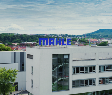 MAHLE Firmengebäude mit blauem Logo, im Hintergrund Stadt, Bäume und Hügel bei Tageslicht.