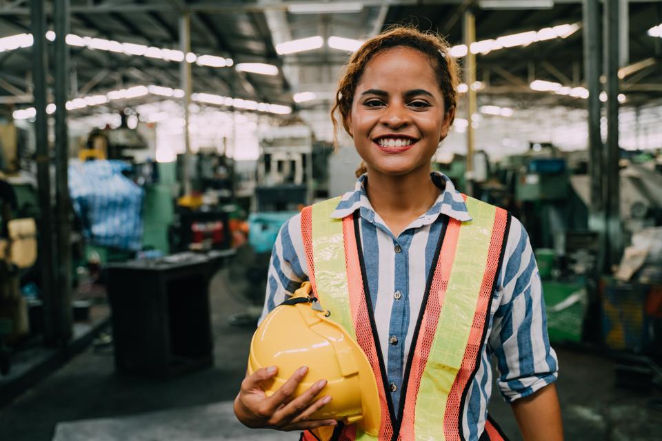 Smiling warehouse worker in hi-vis vest holding hard hat, standing in an industrial factory setting.