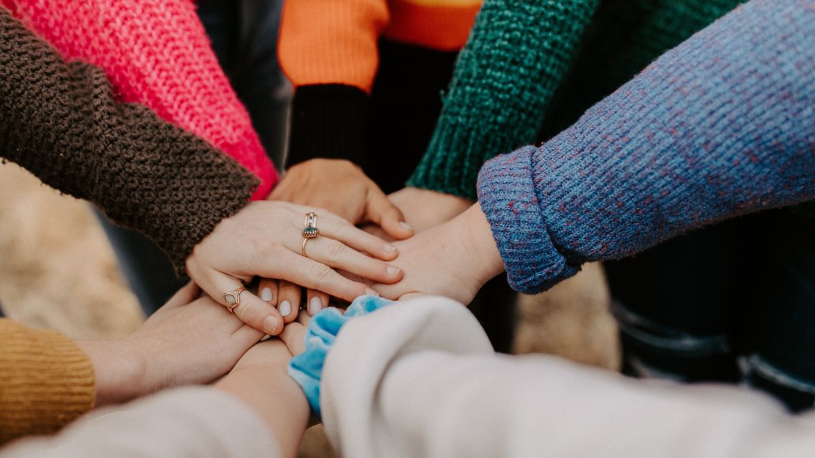 Hands stacked in a circle, wearing colorful sweaters, symbolizing unity.