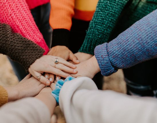 Several people in colorful sweaters join hands in a circle, symbolizing teamwork and unity.