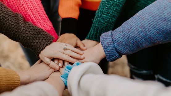 Several people in colorful sweaters join hands in a circle, symbolizing teamwork and unity.