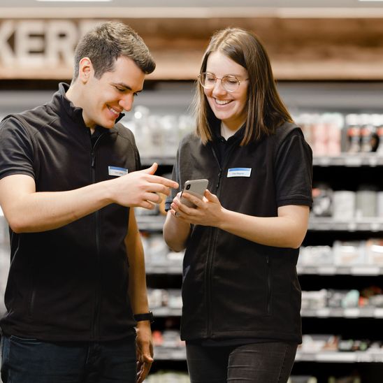 Retail employees in uniform smiling and talking while using a smartphone in a store aisle.