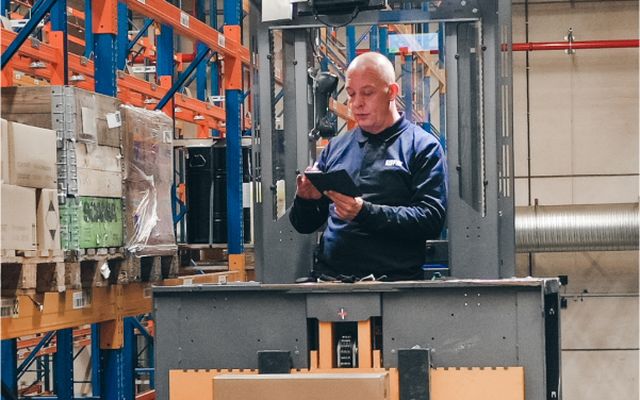 Warehouse worker using a tablet among shelves, no visible UI; environment is industrial.