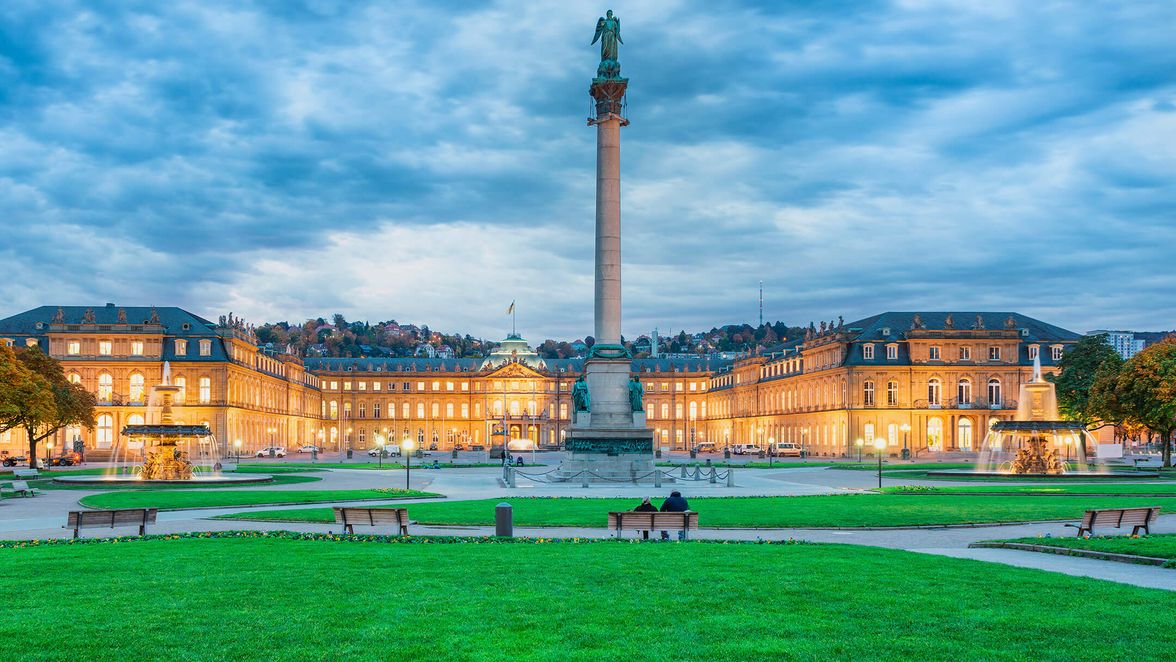 Stuttgart Schlossplatz with illuminated palace, fountains, green lawn, benches, and cloudy sky.