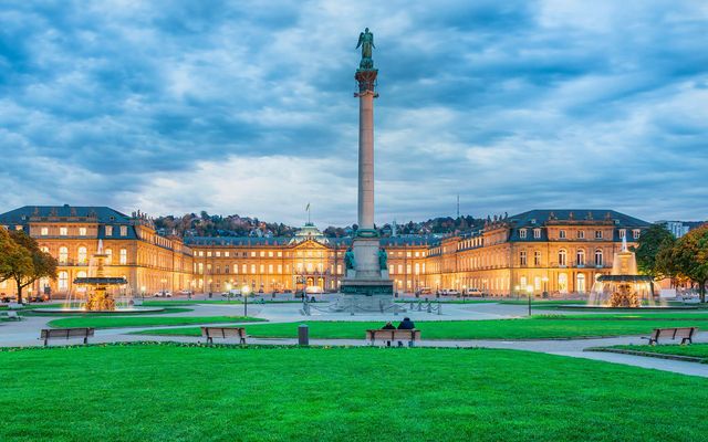 Stuttgart Schlossplatz with illuminated palace, fountains, green lawn, benches, and cloudy sky.
