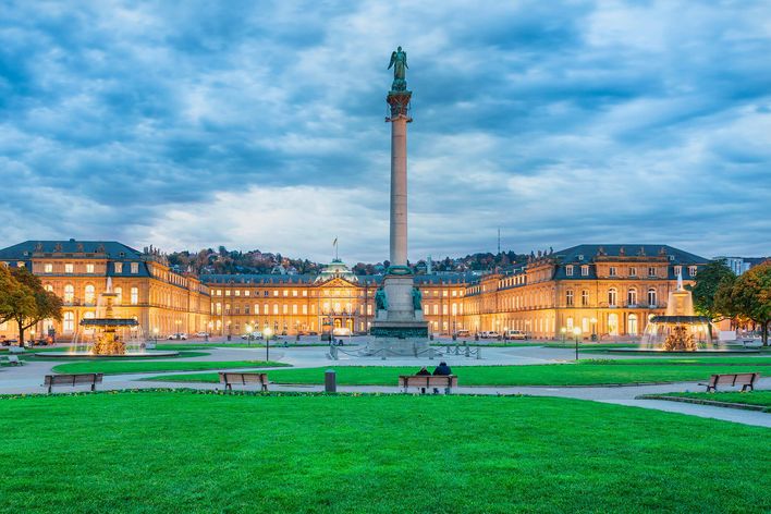 Stuttgart Schlossplatz with illuminated palace, fountains, green lawn, benches, and cloudy sky.
