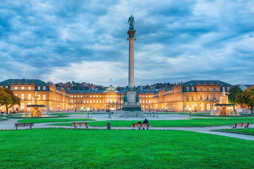 Stuttgart Schlossplatz with illuminated palace, fountains, green lawn, benches, and cloudy sky.