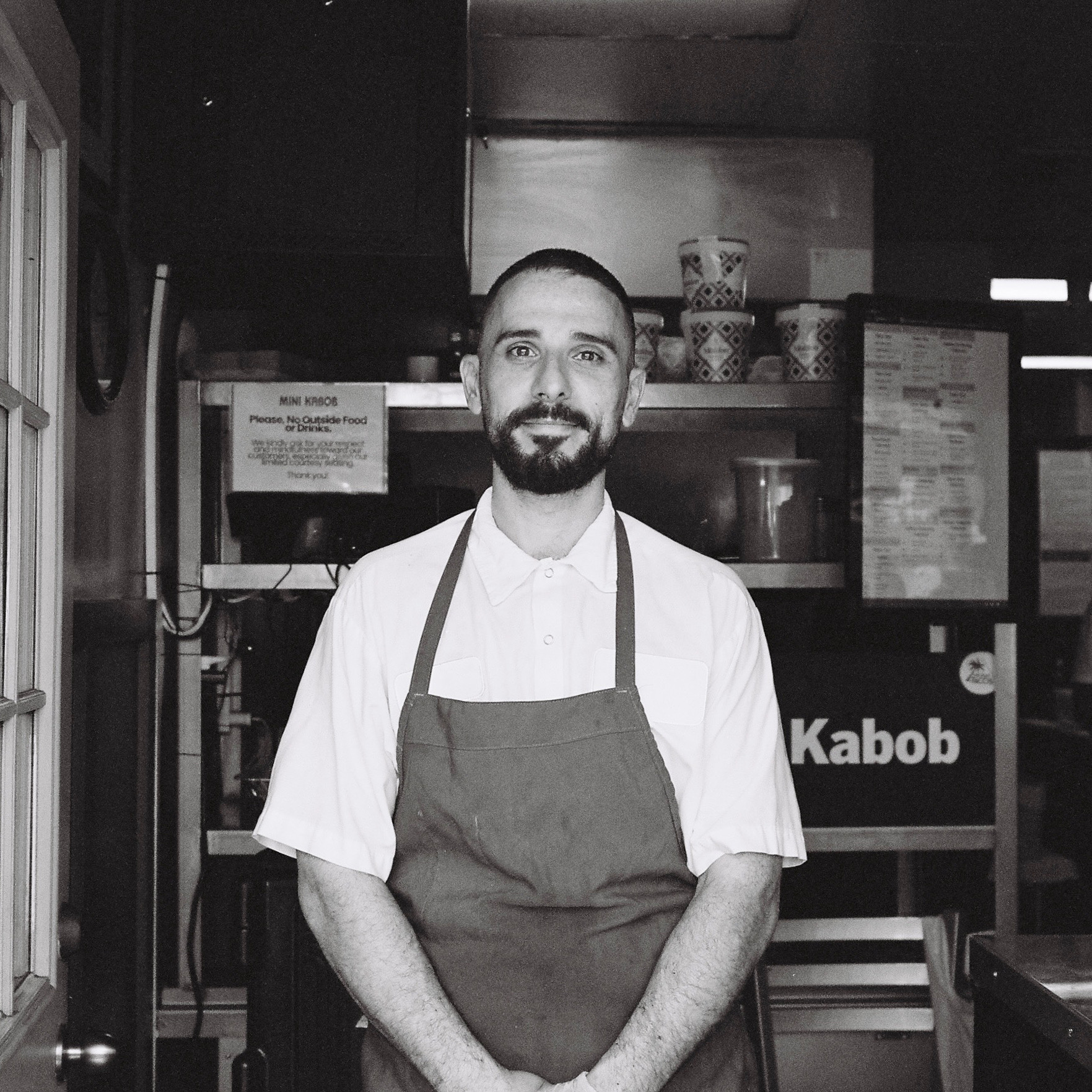 Armen Martirosyan wearing a white shirt and apron stands in a kitchen with menus and cups in the background. Black and white photo.