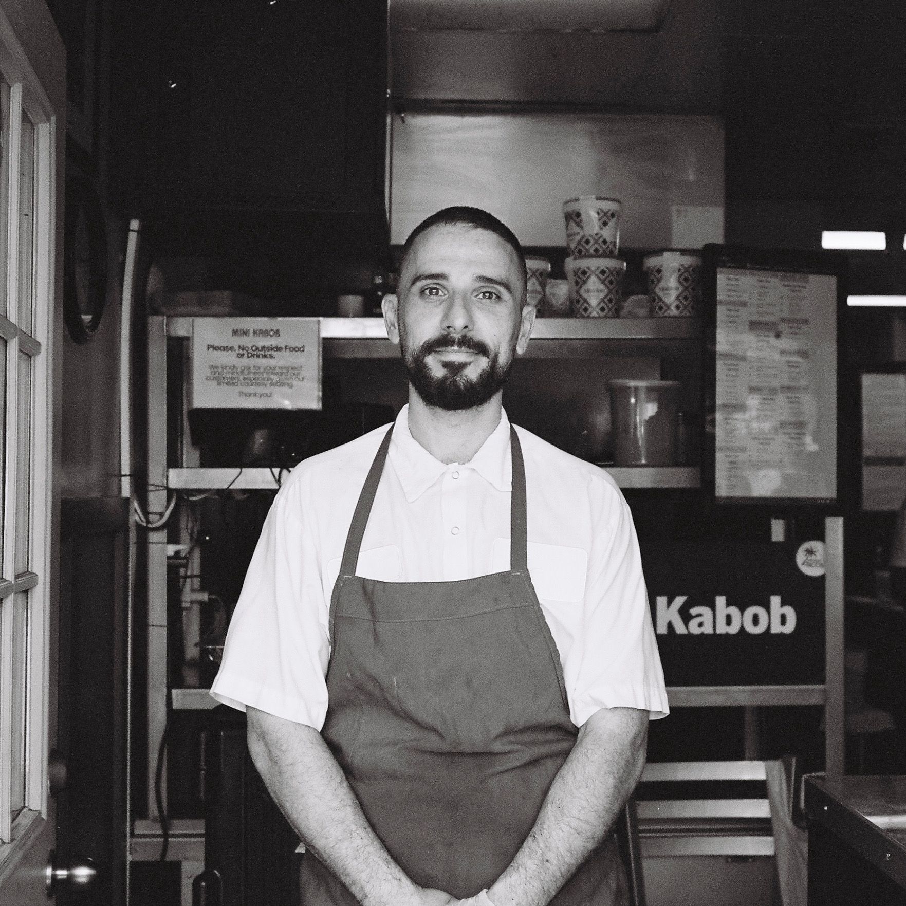 Armen Martirosyan wearing a white shirt and apron stands in a kitchen with menus and cups in the background. Black and white photo.