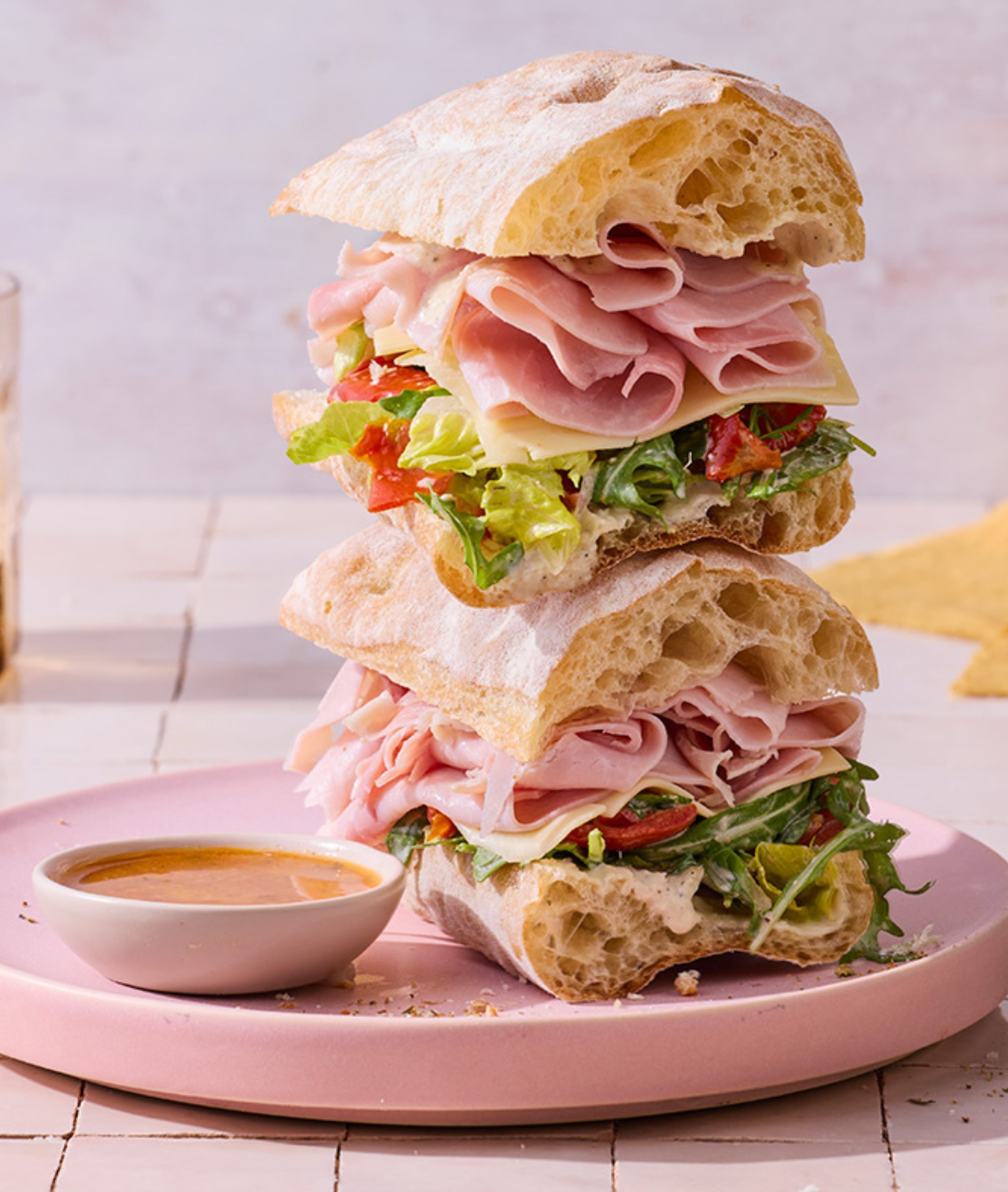 Stacked ciabatta sandwiches with ham, cheese, lettuce, and tomatoes on a pink plate, next to a small bowl of dipping sauce.