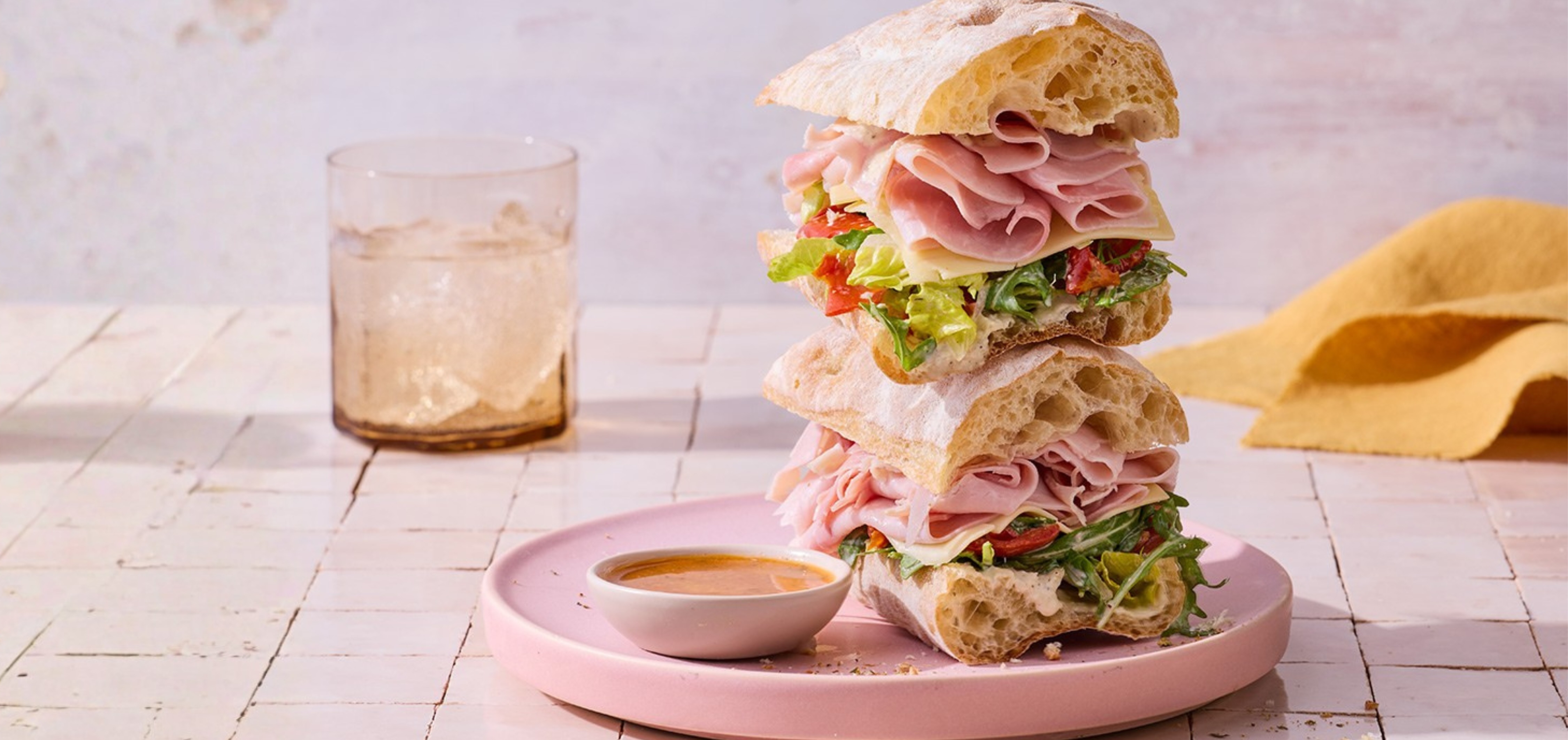 Stacked sandwich with ham, cheese, lettuce, and tomato on ciabatta bread, served on a pink plate with sauce, next to a glass with ice.