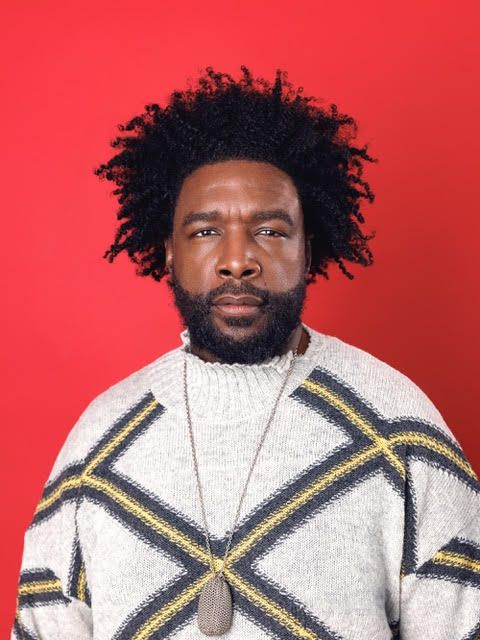 Man with curly hair and beard wearing a patterned sweater and pendant, standing against a red background.